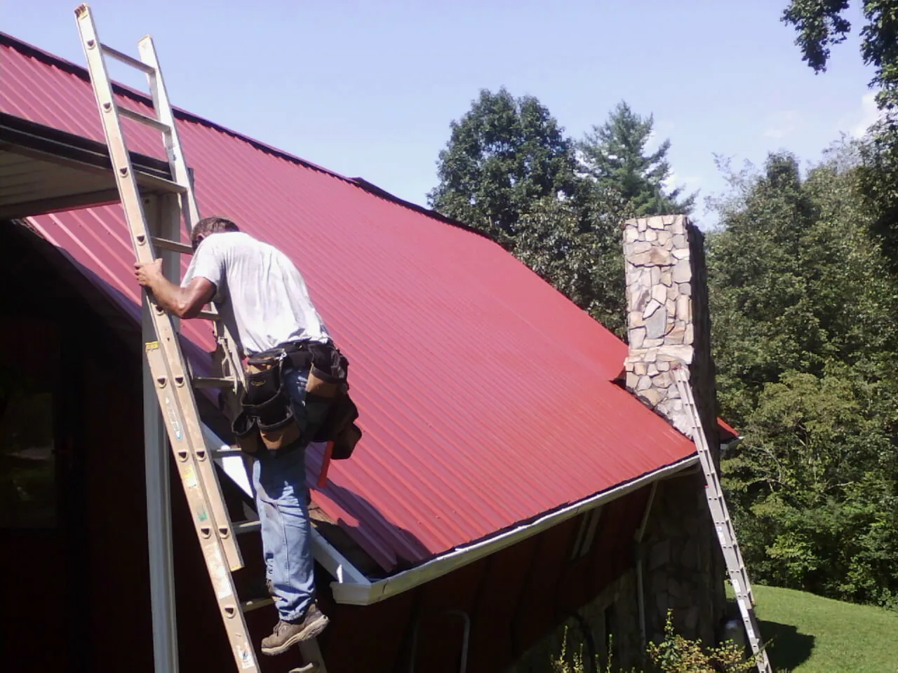 Roofer on a ladder beside a red metal roof and stone chimney — Soffit & Fascia Repair by Roofing Brothers in Giltner